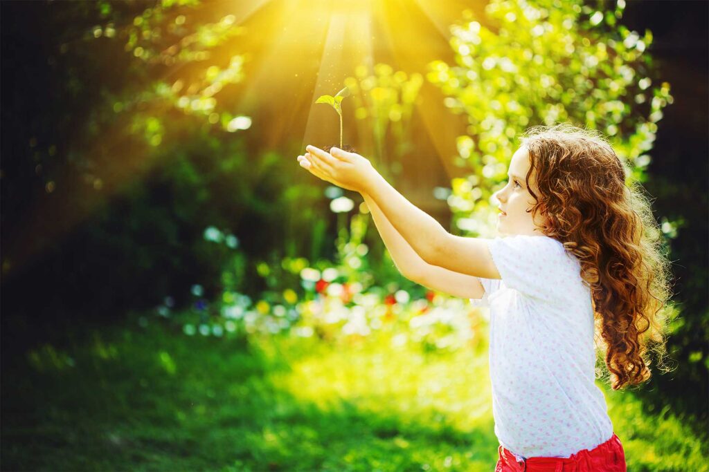 Young girl with her hands outreached holding a small green plant while the sun shines behind her