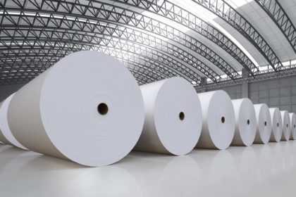 A line of immense rolls of paper sit on the floor of an industrial building.