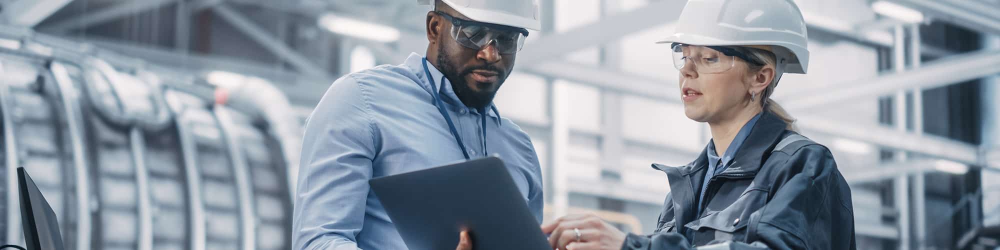 Two coworkers wearing personal protective equipment look at a laptop in a manufacturing facility.