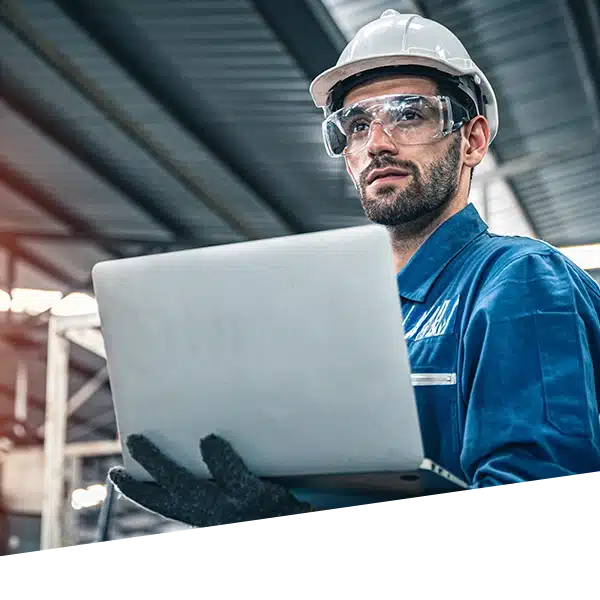 A worker wearing personal protective equipment in an industrial building holds a laptop and looks in the distance.